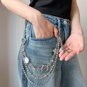 Puede incluir: Cadena de reloj plateada con tres pequeños relojes analógicos y cadenas decorativas. La cadena está sujeta a unos vaqueros azules. Los relojes tienen esferas blancas y negras. La cadena también tiene pequeños dijes de dados.