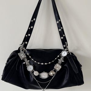 May include: Black leather shoulder bag with silver-toned watch embellishments. The bag features a long strap with silver grommets and a zipper closure. The watch accents include various shapes and sizes, with a heart-shaped pendant.