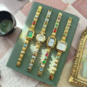 Puede incluir: Cuatro relojes de pulsera dorados con correas decorativas y varias esferas, incluyendo verde, blanco y crema. Las correas están adornadas con gemas y abalorios de colores. Los relojes se exhiben sobre una superficie de cuero verde.