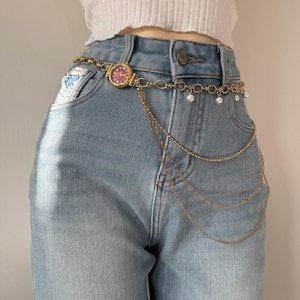 Peut inclure: Ceinture chaîne dorée avec une petite montre à cadran rouge. La ceinture comporte plusieurs brins de chaînes délicates et de fausses perles, portée sur un jean en denim bleu clair.