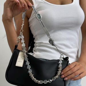 May include: Black baguette handbag with a silver watch-style strap and a beaded accent. The strap features a watch face and clear, faceted beads. The bag has a small tag attached.