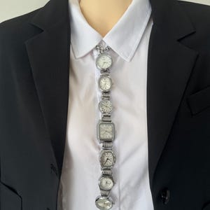 Puede incluir: Un collar de reloj plateado único compuesto por varios relojes pequeños con esferas blancas y correas plateadas. El collar se lleva sobre una camisa blanca y una blazer negra.