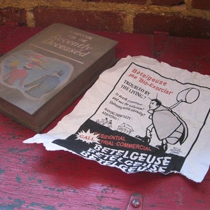 K&ouml;nnte beinhalten: Eine braune Box mit dem Text "Handbook for the Recently Deceased" und einem Cartoon-Bild einer Person in einem rot-blauen Outfit. Ein wei&szlig;es Papier mit schwarzem Text und einem Cartoon-Bild einer Person, die auf einem K&auml;fer reitet, mit dem Text "Betelgeuse the 'Bio-Exorcist'" liegt auf einer roten Holzoberfl&auml;che.