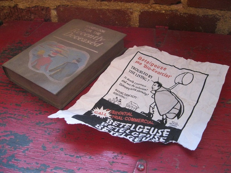 K&ouml;nnte beinhalten: Eine braune Box mit dem Text "Handbook for the Recently Deceased" und einem Cartoon-Bild einer Person in einem rot-blauen Outfit. Ein wei&szlig;es Papier mit schwarzem Text und einem Cartoon-Bild einer Person, die auf einem K&auml;fer reitet, mit dem Text "Betelgeuse the 'Bio-Exorcist'" liegt auf einer roten Holzoberfl&auml;che.