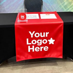 May include: A woman stands near a black table with a red runner that says "Your Logo Here" in white. The background is a colorful geometric design. Plants are on shelves and a blue cooler is to the right.