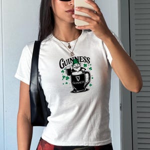 May include: White t-shirt featuring a Guinness design. The graphic includes the Guinness logo, a cartoon gnome holding a pint of beer, and green shamrocks. The shirt is a crew neck style.