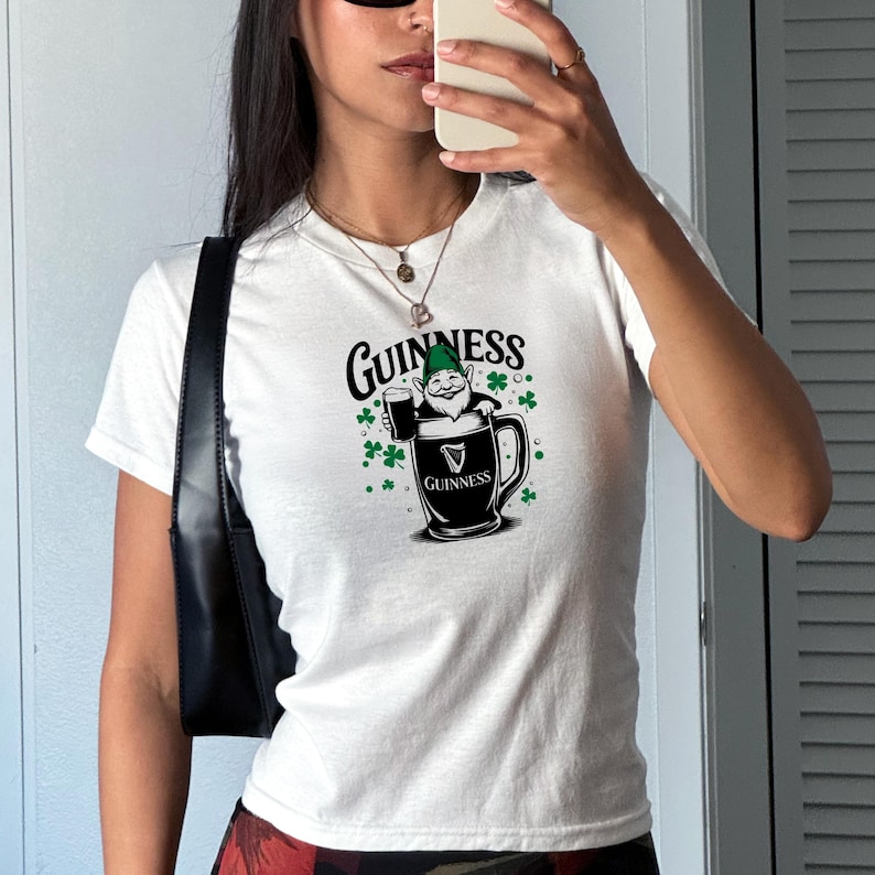 May include: White t-shirt featuring a Guinness design. The graphic includes the Guinness logo, a cartoon gnome holding a pint of beer, and green shamrocks. The shirt is a crew neck style.