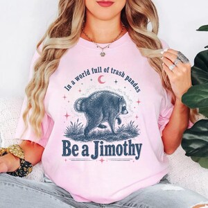 May include: A light pink t-shirt featuring a dark blue graphic of a raccoon walking through grass. Above the raccoon, the text reads In a world full of trash pandas, and below it, the text reads Be a Jimothy, accented by a small red crescent moon.