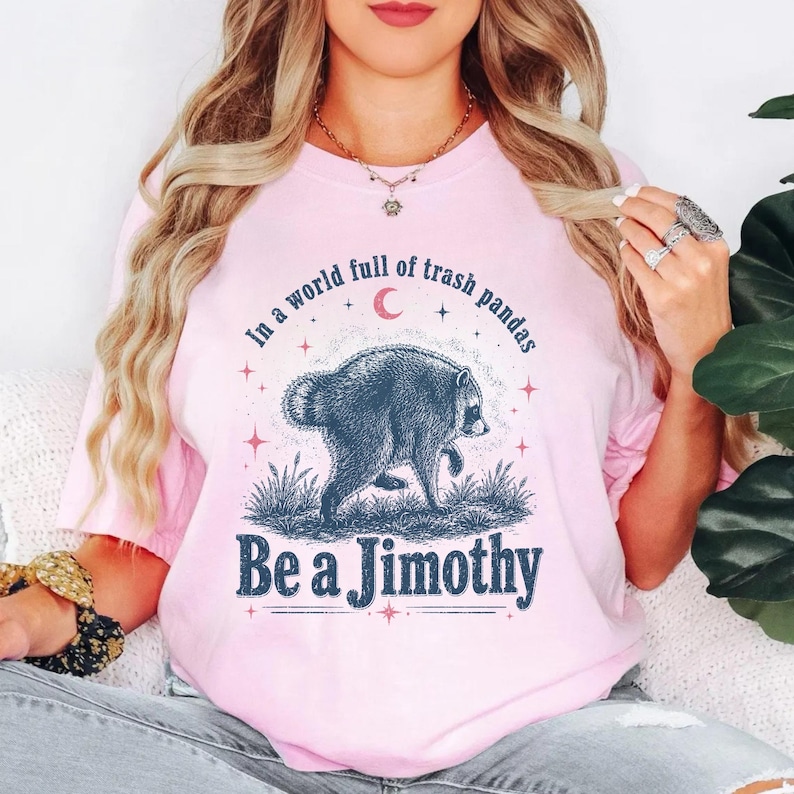 May include: A light pink t-shirt featuring a dark blue graphic of a raccoon walking through grass. Above the raccoon, the text reads In a world full of trash pandas, and below it, the text reads Be a Jimothy, accented by a small red crescent moon.