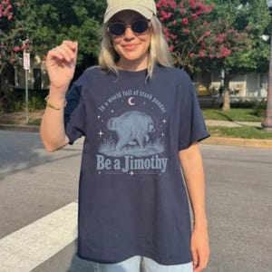 May include: A navy blue t-shirt featuring a graphic of a raccoon in a grassy field under a crescent moon and stars. The text reads, In a world full of trash pandas Be a Jimothy, printed in a light blue font with decorative line accents.
