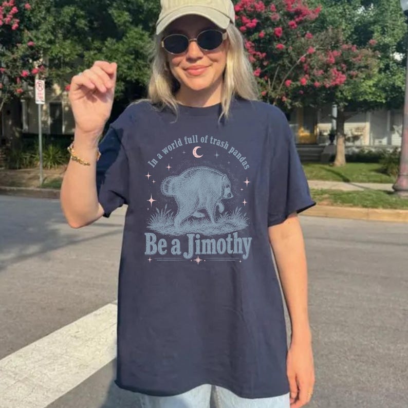 May include: A navy blue t-shirt featuring a graphic of a raccoon in a grassy field under a crescent moon and stars. The text reads, In a world full of trash pandas Be a Jimothy, printed in a light blue font with decorative line accents.