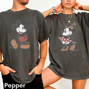 May include: Two dark gray t-shirts featuring Mickey and Minnie Mouse. Mickey is wearing red shorts and brown shoes. Minnie is wearing a red polka dot skirt and brown shoes. The word "Disney" is printed on the shirts.