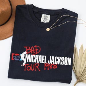 May include: A black crew neck t-shirt featuring a graphic print with the text BAD MICHAEL JACKSON TOUR 1988 in red and white. The design includes a white silhouette of a dancer and a Pepsi logo. The shirt has a Comfort Colors brand neck label.