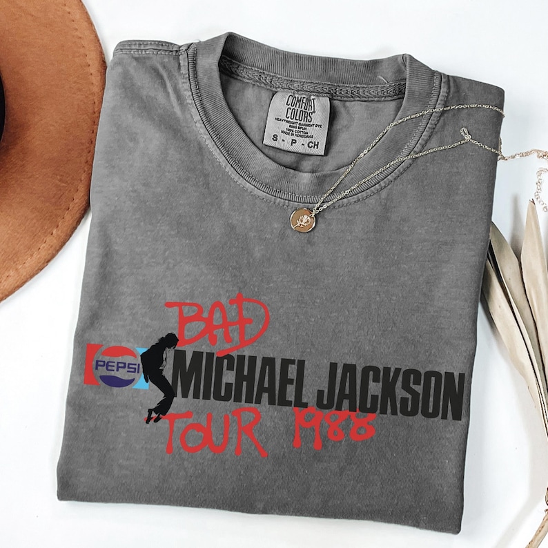 May include: A charcoal gray short-sleeve t-shirt featuring a graphic print with the red text BAD and TOUR 1988, black text MICHAEL JACKSON, a Pepsi logo, and a silhouette of a dancer. The shirt has a Comfort Colors label in the inner neckline.