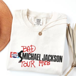 May include: An off-white short-sleeve t-shirt featuring a graphic print with the Pepsi logo, a silhouette of Michael Jackson, and the red and black text "BAD MICHAEL JACKSON TOUR 1988". The shirt has a Comfort Colors brand label at the inner neckline.