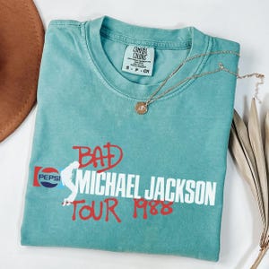 May include: A teal short-sleeve t-shirt with a Comfort Colors label in the neck. The front features a graphic with the text "BAD MICHAEL JACKSON TOUR 1988" in red and white, alongside a Pepsi logo and a white silhouette of a dancing figure.