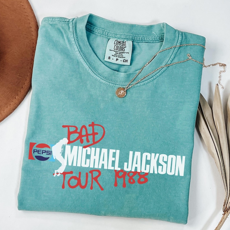May include: A teal short-sleeve t-shirt with a Comfort Colors label in the neck. The front features a graphic with the text "BAD MICHAEL JACKSON TOUR 1988" in red and white, alongside a Pepsi logo and a white silhouette of a dancing figure.