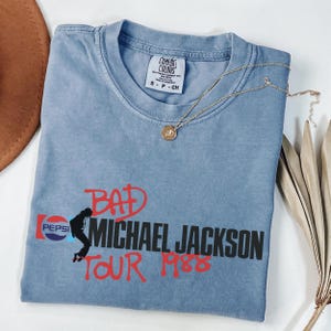 May include: A blue short-sleeved t-shirt featuring a graphic print that includes the Pepsi logo, a silhouette of Michael Jackson, and the text "BAD MICHAEL JACKSON TOUR 1988" in red and black. The shirt has a Comfort Colors brand neck label.