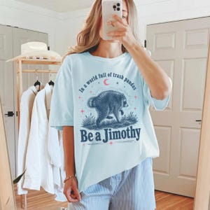 May include: A light blue short-sleeve t-shirt featuring a graphic of a raccoon in grass with a crescent moon and stars. The text reads In a world full of trash pandas Be a Jimothy. The design uses dark blue, red, and white ink colors.