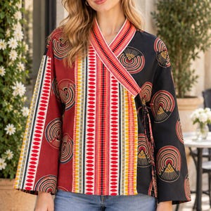 Puede incluir: Una blusa cruzada de manga larga con un diseño de bloques de color, con un lado rojo oscuro y otro negro, ambos adornados con patrones geométricos circulares. El panel central muestra rayas verticales en rojo, naranja, amarillo y blanco.