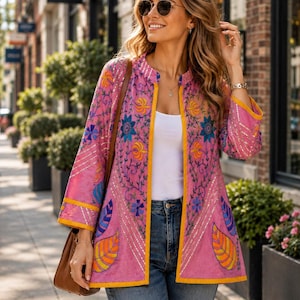 Könnte beinhalten: Eine pinkfarbene, vorne offene Jacke mit bunten Blumen- und Blattstickereien, goldenen Metallic-Fäden und einem einfarbigen gelben Saum am Kragen, an der Vorderseite und an den Ärmelbündchen, getragen über einem weißen Oberteil und blauen Jeans.