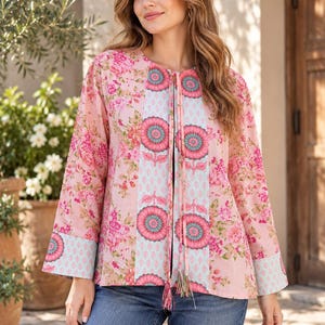 May include: A long-sleeved blouse featuring a light pink base with a floral pattern on the sides and a central vertical panel in light blue with a repeating pink and teal circular flower design, finished with a tie-front closure and tassels.