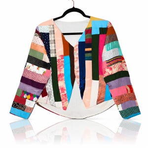 May include: A long-sleeved jacket constructed from a patchwork of various fabric strips in diverse colors and patterns, including floral, polka dot, and solid textures. The garment features a V-neckline, a front button closure, and a white interior lining.