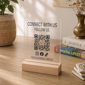 May include: A clear acrylic sign with a rectangular wooden base featuring the black text CONNECT WITH US FOLLOW US, a central QR code with a thumbs-up icon, social media icons for Instagram, Facebook, and TikTok, and the text YOUR BUSINESS HANDLE.