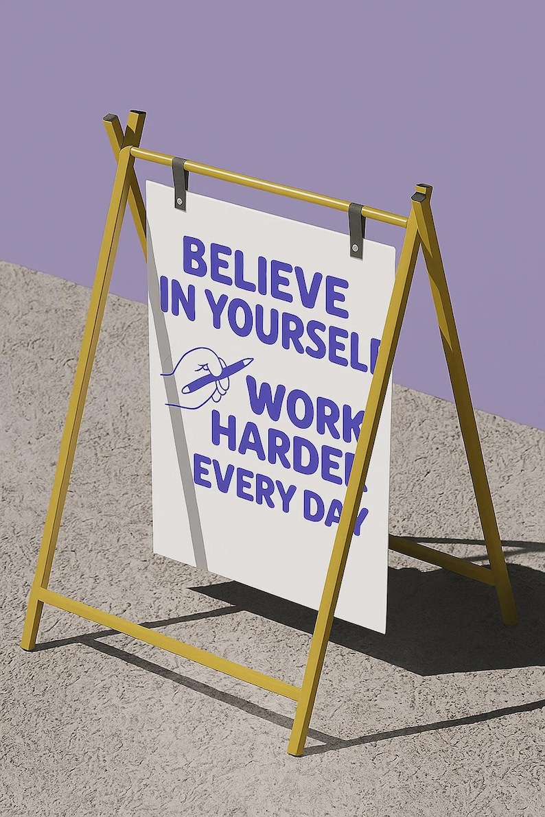 May include: A-frame sign with a yellow frame and a white sign. The sign has the text "BELIEVE IN YOURSELF! WORK HARDER EVERY DAY" in blue. A drawing of a hand holding a pen is also on the sign.