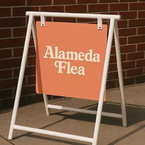 May include: A white metal A-frame sign with an orange sign face. The sign reads "Alameda Flea" in white, stylized lettering. The sign is set on a concrete surface in front of a brick wall.