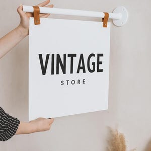 Peut inclure: Une enseigne blanche avec les mots "VINTAGE STORE" en lettres noires est suspendue &agrave; une barre blanche avec des lani&egrave;res en cuir marron. L'enseigne est tenue par une personne. L'enseigne est fix&eacute;e sur un mur blanc.