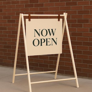 May include: A-frame sign with the words "NOW OPEN" in dark green lettering. The sign is cream-colored and hangs from a light wood frame with brown leather straps. The background is a brick wall.