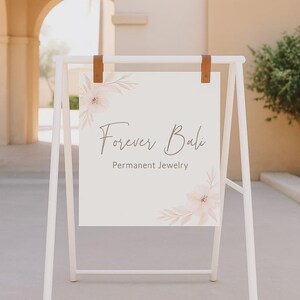 May include: A white A-frame sign with the text "Forever Bali Permanent Jewelry." The sign is held by brown leather straps and features floral accents in the corners. The sign is outdoors, with a building and greenery in the background.