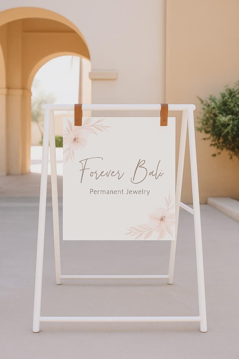 May include: A white A-frame sign with the text "Forever Bali Permanent Jewelry." The sign is held by brown leather straps and features floral accents in the corners. The sign is outdoors, with a building and greenery in the background.