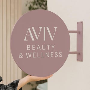 Peut inclure: Un panneau rond mauve avec le texte "AVIV BEAUTY & WELLNESS" en lettres blanches. Le panneau est mont&eacute; sur un mur avec un support mauve assorti. Le panneau est tenu par une personne.