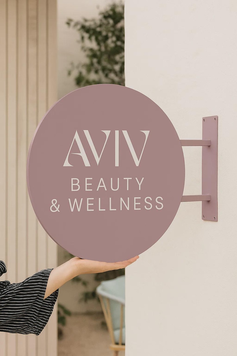 Peut inclure: Un panneau rond mauve avec le texte "AVIV BEAUTY & WELLNESS" en lettres blanches. Le panneau est mont&eacute; sur un mur avec un support mauve assorti. Le panneau est tenu par une personne.