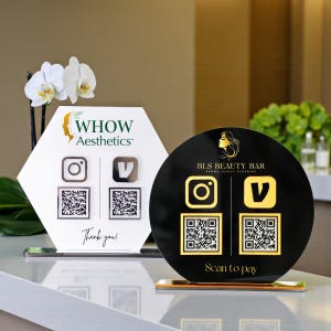 May include: A white hexagonal sign with the text WHOW Aesthetics and two QR codes, alongside a black circular sign with the text BLS BEAUTY BAR, Scan to pay, and two gold QR codes. Both signs feature social media icons and stand on flat bases.