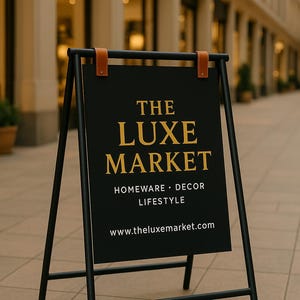 May include: Black metal A-frame sign with the text "THE LUXE MARKET" in gold lettering. The sign also includes "HOMEWARE &bull; DECOR &bull; LIFESTYLE" and a website address. Brown leather straps secure the sign.