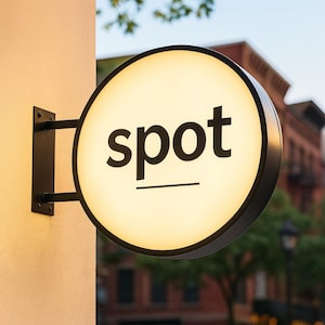 May include: A round, illuminated sign with the word "spot" in black, sans-serif font. The sign is mounted on a wall with a black metal bracket. The sign has a black border and a cream-colored illuminated face.