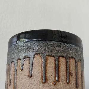 Könnte beinhalten: Ein zylindrischer Keramikbecher mit gesprenkelter beiger Basis und einem dunkel glänzenden schwarzen Rand. Der obere Teil weist eine texturierte, metallisch wirkende Glasur auf, die über die matte beige Oberfläche läuft.