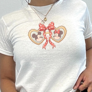Op de afbeelding: Wit T-shirt met een Mickey en Minnie Mouse ontwerp. De afbeelding bevat hartvormige kaders met de gezichten van de personages, een rode strik met stippen en linten. Het T-shirt heeft korte mouwen en is gemaakt van zacht materiaal.