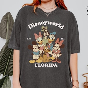 May include: Dark gray t-shirt featuring a vintage-style graphic of Disney characters, including Mickey and Minnie Mouse, Donald and Daisy Duck, and Goofy. The text "Disneyworld Florida Est. 1971" is printed above and below the characters.