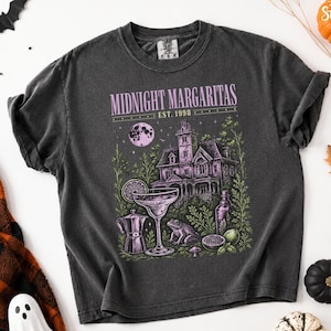 May include: A charcoal gray short-sleeve t-shirt featuring a graphic print with the text "MIDNIGHT MARGARITAS EST. 1998". The design includes a large house, a margarita glass, a frog, a coffee pot, a bottle, and a full moon in purple and green.