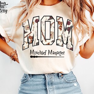Peut inclure: Un t-shirt à manches courtes couleur ivoire arborant le mot MOM en grandes lettres majuscules remplies d'un motif d'icônes sur le thème des sorciers. Sous le mot MOM, le texte indique Mischief Manager avec un graphique de baguette noire.