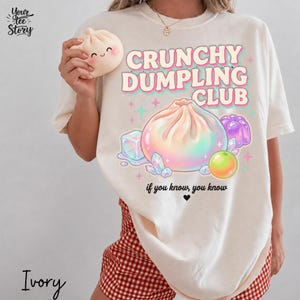 以下が含まれることがあります： 「CRUNCHY DUMPLING CLUB」と「if you know, you know」の文字、小さなハートが描かれたアイボリーのTシャツ。デザインには、大きな虹色の餃子、紫色のゼリー、氷のキューブ、小さな黄色い球体がプリントされています。