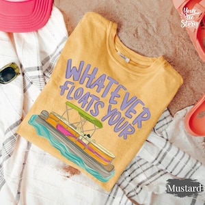 May include: Mustard-colored t-shirt with the text "Whatever Floats Your" and a graphic of a pontoon boat. The boat has orange, pink, and green accents. The shirt is displayed on a white and gray striped towel with a pink hat and sunglasses.