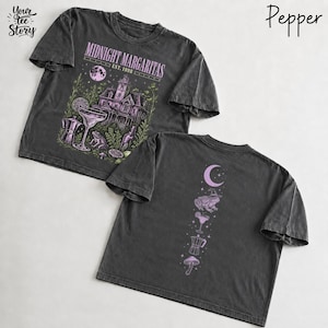 May include: A charcoal gray t-shirt featuring a front graphic with a house, a cocktail glass, a frog, and the text Midnight Margaritas Est. 1998. The back graphic displays a vertical arrangement of a crescent moon, a frog, a cocktail, and a mushroom.