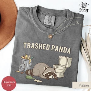 May include: A grey t-shirt featuring the words "TRASHED PANDA" and a cartoon raccoon next to a bin and toilet. The shirt is a Comfort Colors brand, size S-P-CH.
