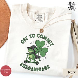 Może przedstawiać: Kremowa koszulka z zieloną żabą na skuterze, w kapeluszu i pelerynie czarownicy oraz dużym koniczyną. Tekst brzmi "OFF TO COMMIT SHENANIGANS" w kolorze zielonym. Złoty naszyjnik jest zarzucony na koszulkę.