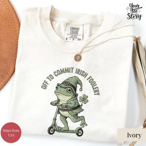 May include: Ivory-colored t-shirt with a frog riding a scooter, wearing a leprechaun hat and cloak. Text reads "OFF TO COMMIT IRISH FOOLERY." A gold necklace is draped across the shirt. The shirt is labeled "Comfort Colors."